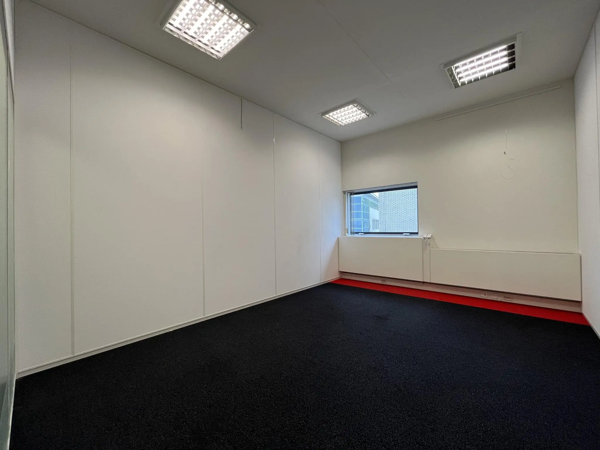 Leeg kantoor- of praktijkruimte in Leidse Rijn met witte muren, zwart tapijt en een klein raam.